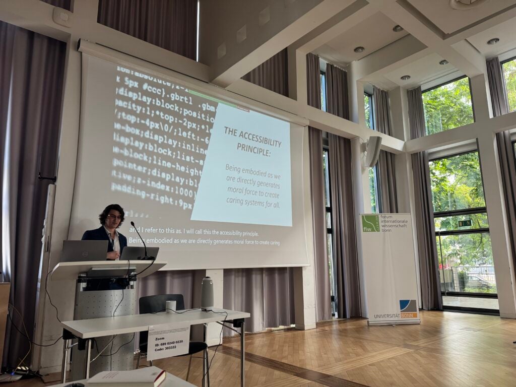 Professor Joel Reynolds stands at a podium in front of a screen with a slide reading "The Accessibility Principle" in a room with floor to ceiling windows at Universitat Bonn